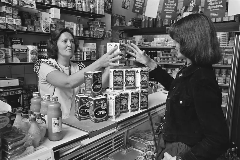 Een winkelierster geeft een klant over de toonbank pakken koffie van Van Nelle en Douwe Egberts.