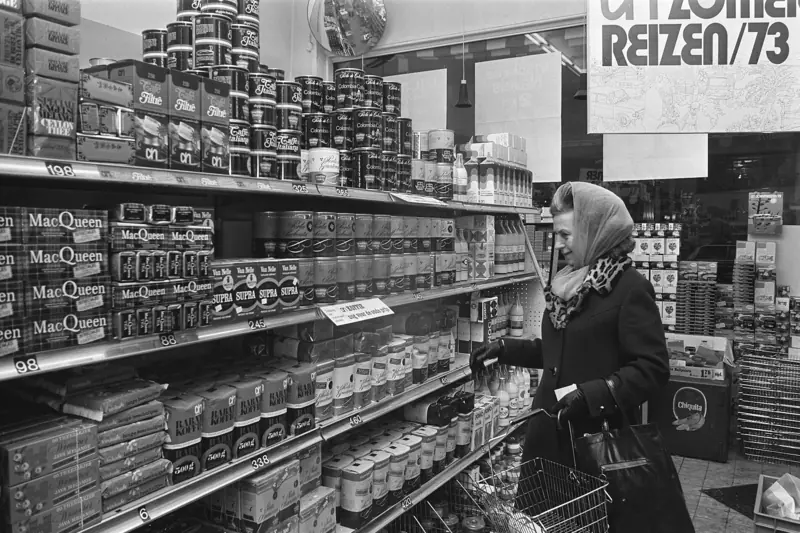 Een vrouw met hoofddoek en boodschappenmandje pakt koffie uit een supermarktschap. Onder elk pak staat een prijskaartje.