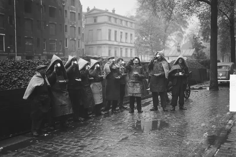 Een rij kolenmannen met zakken als capuchon over hun hoofd drinkt koffie op een natte straat.
